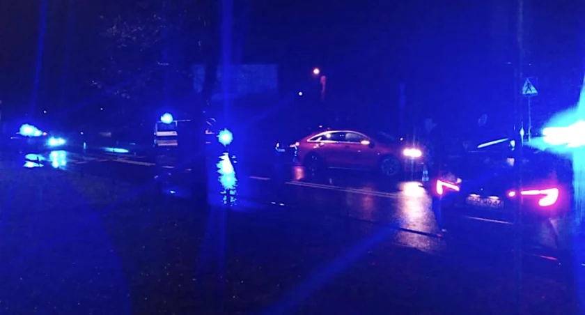 Zdjęcie przedstawia auto osobowe i radiowozy z włączonymi sygnałami świetlnymi.